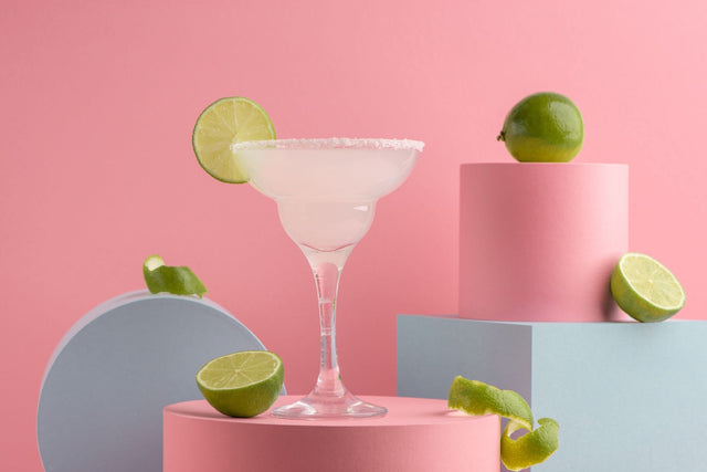 Canna-Rita THC margarita in a salt-rimmed margarita glass surrounded by fresh limes on pink and blue risers