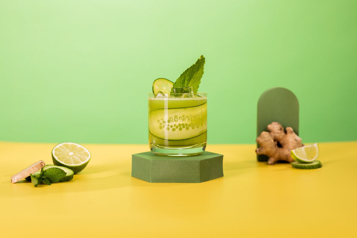 Daily Greens THC cocktail in a rocks glass garnished with cucumber and mint, with lime and fresh ginger on a green backdrop