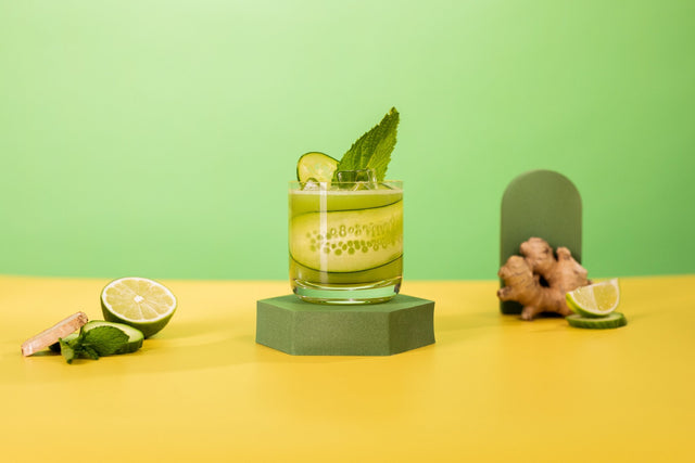 Daily Greens THC cocktail in a rocks glass garnished with cucumber and mint, with lime and fresh ginger on a green backdrop