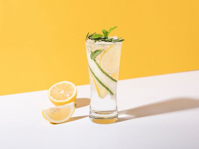 Flora Highball THC cocktail in a tall glass over ice with cucumber ribbons, lemon wheels and mint, on a yellow backdrop