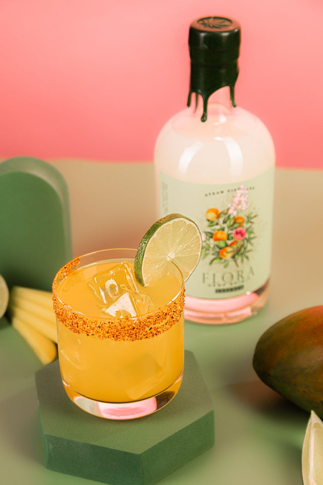Mango Flora-Rita THC cocktail in a Tajin-rimmed rocks glass with a lime wheel, beside a FLORA spirit bottle and fresh mango