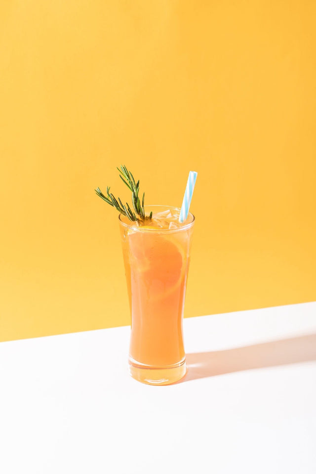 Orange Crush THC cocktail in a tall collins glass, garnished with a rosemary sprig and an orange slice, against a yellow backdrop
