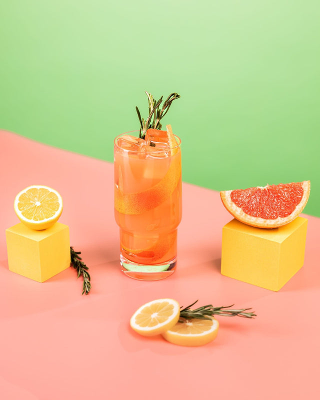 Flora Paloma THC cocktail over ice garnished with rosemary, surrounded by grapefruit and lemon slices on a pink and green backdrop