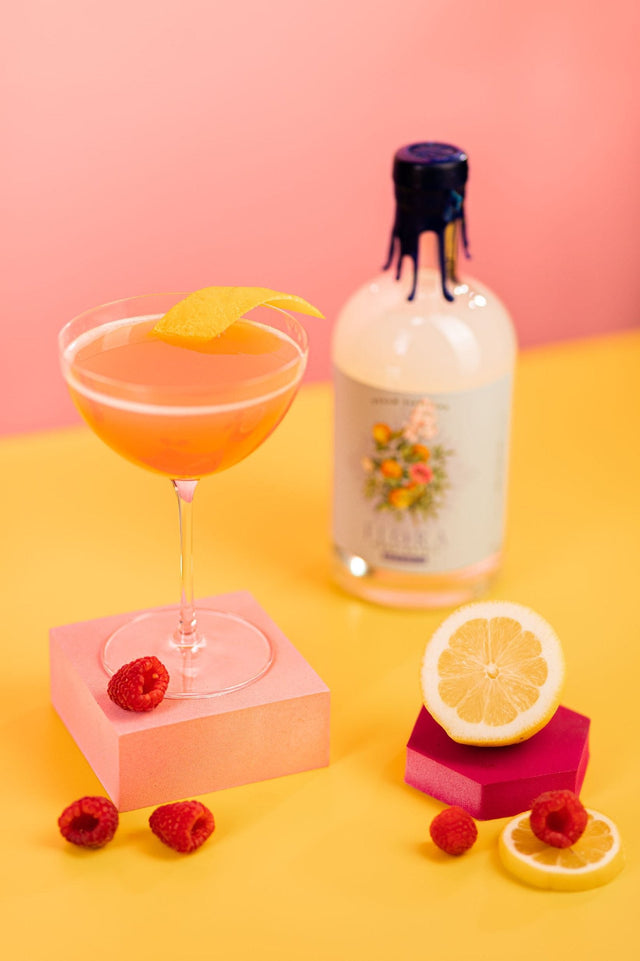 Raspberry Chill THC cocktail in a coupe glass with a foam top and citrus twist, beside a bottle of FLORA spirit and fresh raspberries