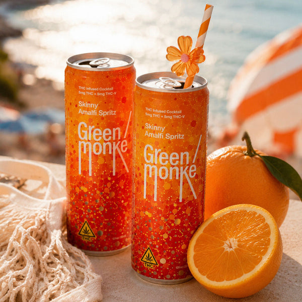 Two cans of Green Monke Skinny Amalfi Spritz THC & THCV cocktails are displayed on a beach table with oranges, a net, and a striped umbrella in the background.